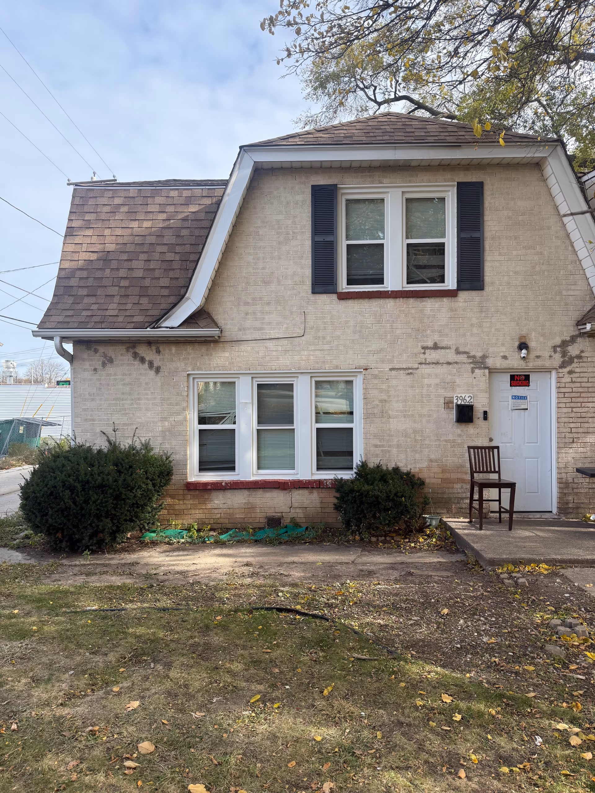 Front exterior of a beige brick two-story home with shutters and a chair by the door.