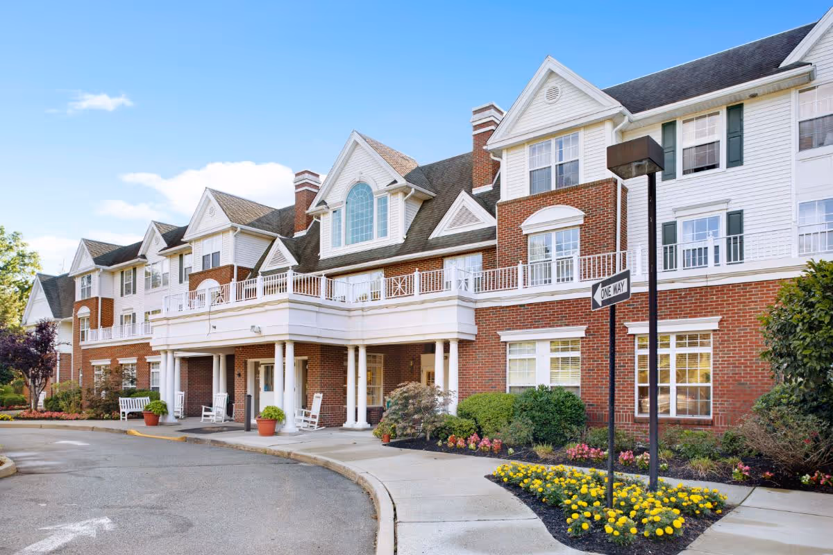Front entrance of a multi-story brick and white senior living facility with columns, landscaping, and a 'One Way' sign.