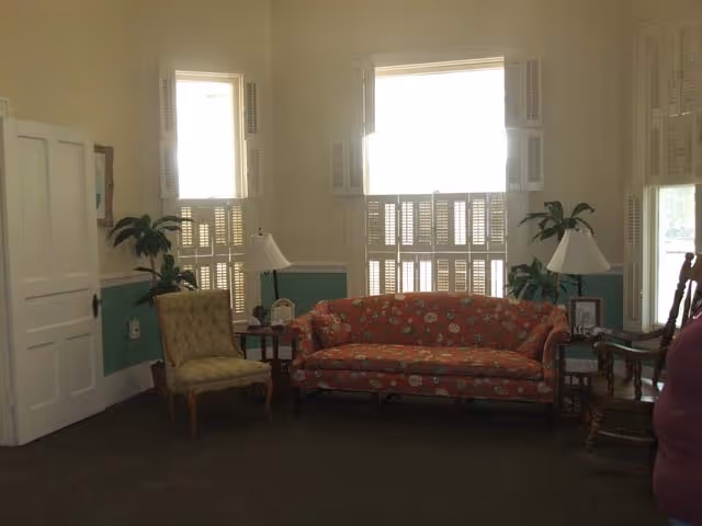 A living room with a floral patterned sofa, a cushioned armchair, two side tables with lamps, and several potted plants. The room has three windows with white shutters and light green walls with white trim.