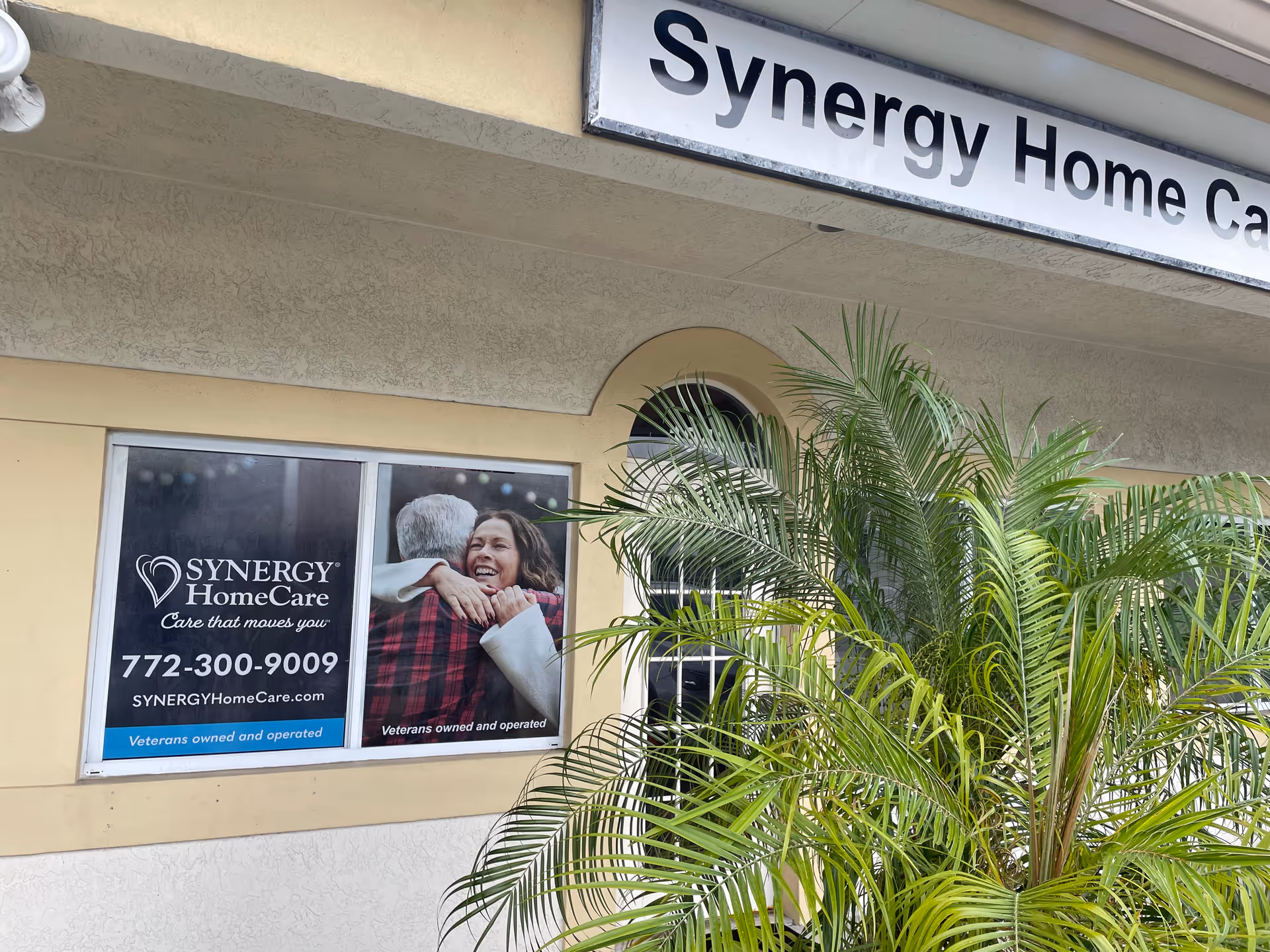 Exterior view of Synergy HomeCare building with signage, windows, and a large palm plant.