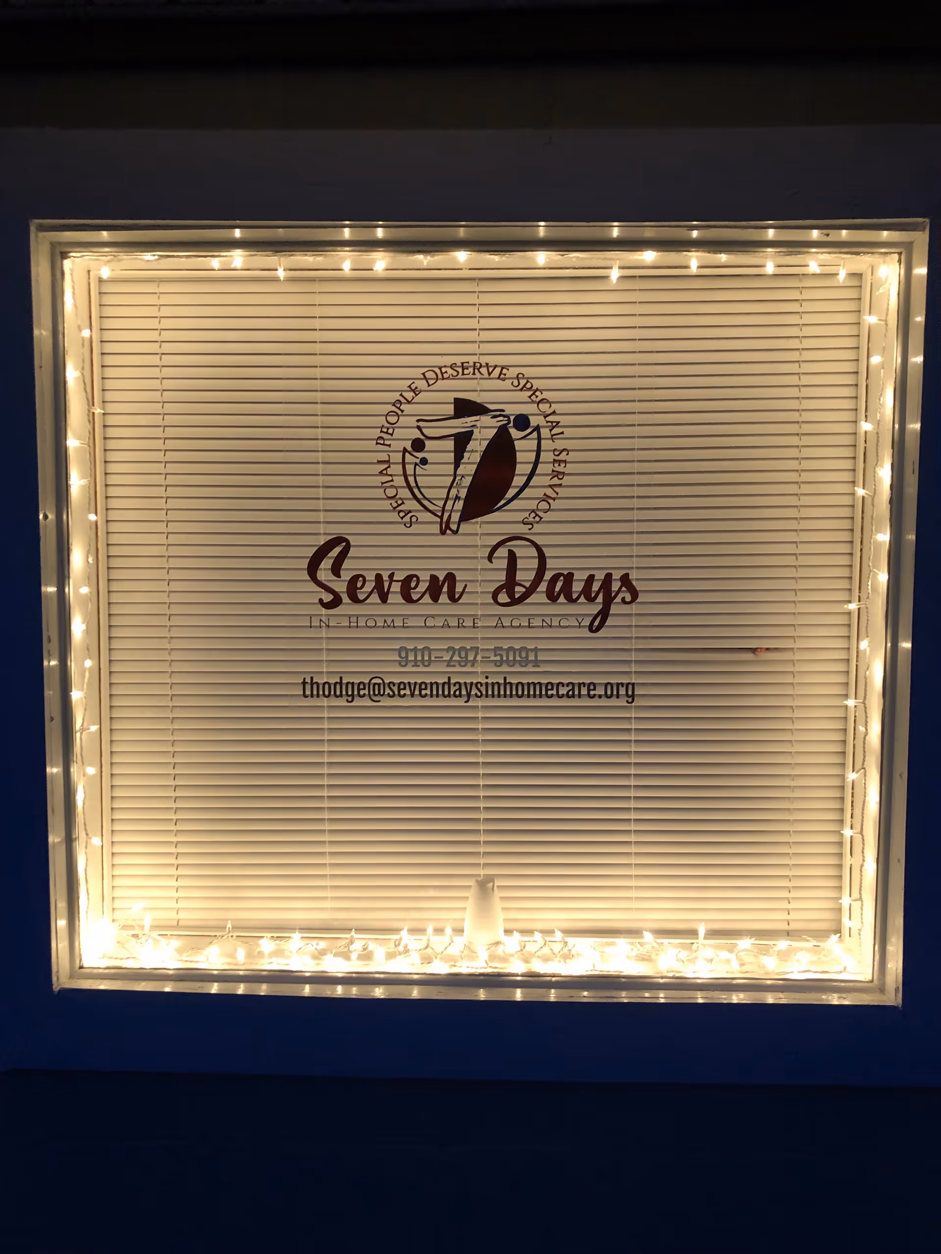 Window sign for Seven Days In-Home Care Agency with blinds and string lights.
