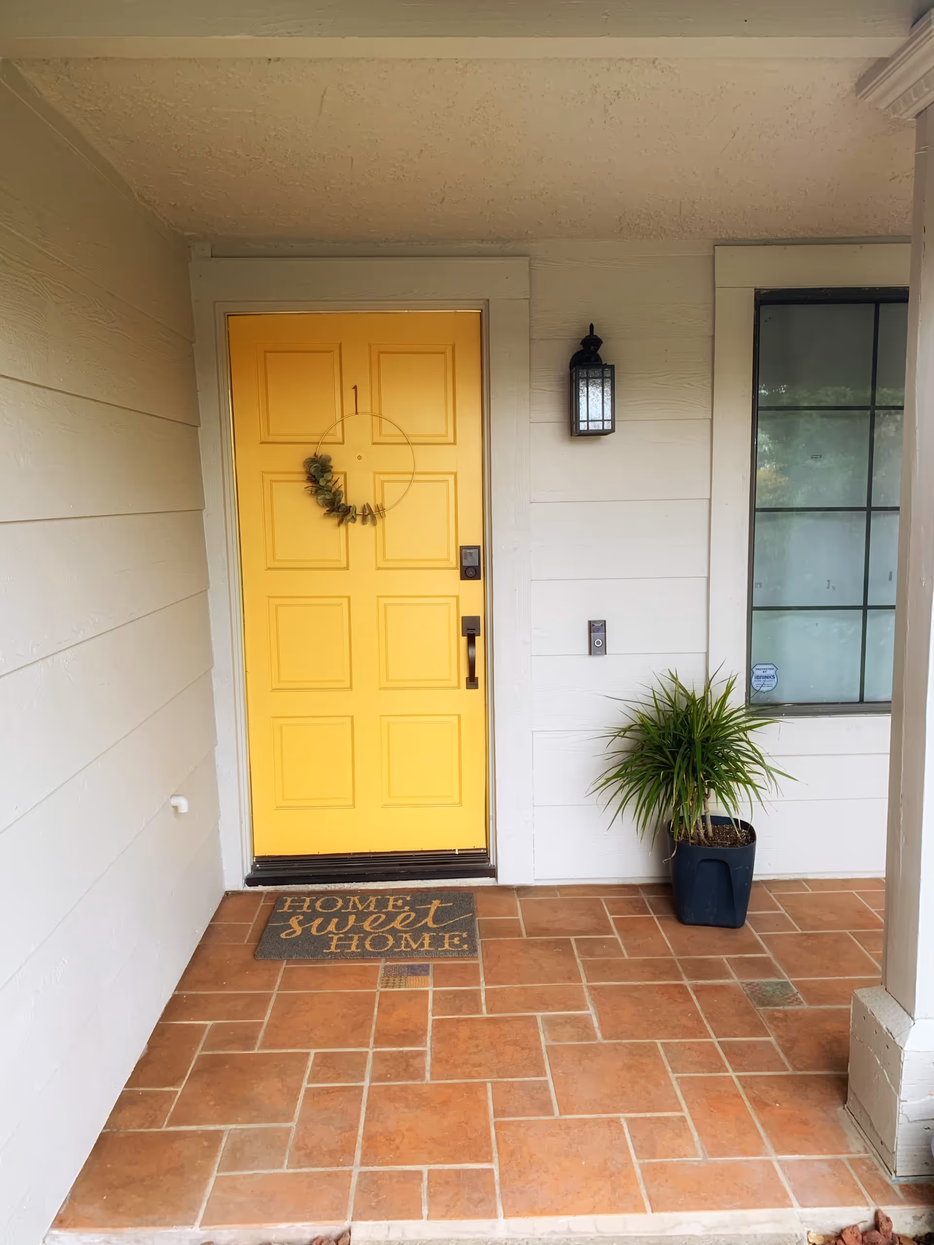 Front porch area with a bright yellow door decorated with a circular wreath. A welcome mat in front of the door reads 'Home sweet Home.' There is a black wall-mounted lantern light fixture to the right of the door and a potted green plant next to a window with black framing. The floor is tiled with reddish-brown tiles.