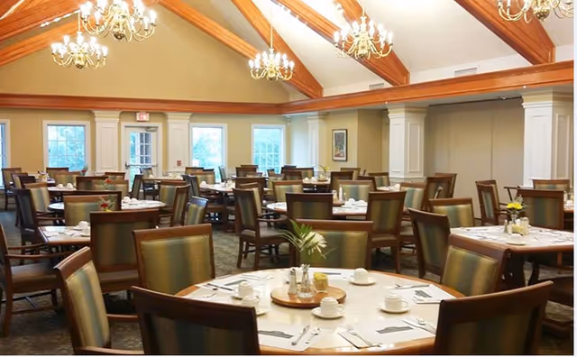 An elegant senior dining room with round tables, chairs, chandeliers, and wooden vaulted beams.