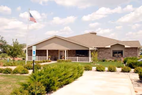 Single-story senior living facility exterior with walkway, landscaping, and an American flag