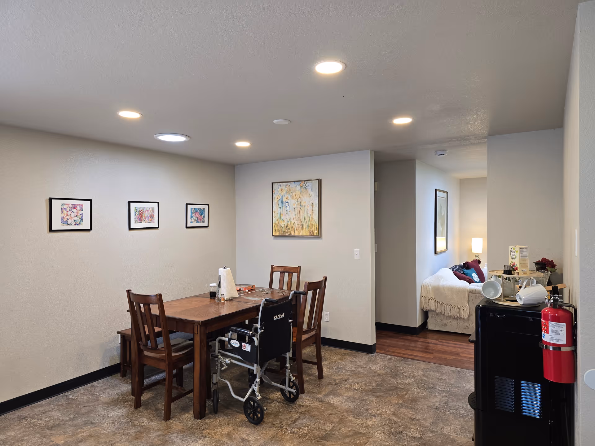 Dining area with wooden table, chairs, a wheelchair, and a doorway to a bedroom.