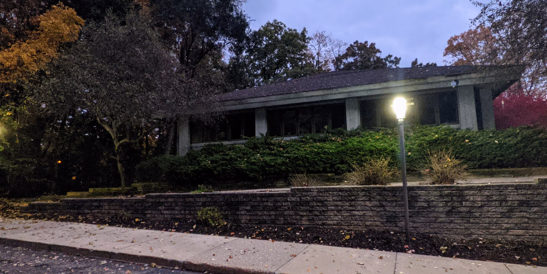 Exterior view of a low, single-story building behind a hedge with a lit street lamp.