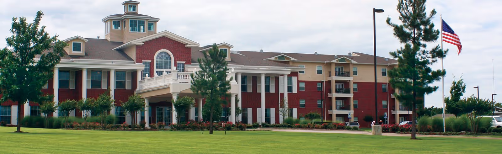 Exterior view of Meadow Lake Senior Living facility showing a large multi-story building with red and beige walls, white columns at the entrance, surrounded by green lawn, trees, and an American flag on a flagpole.