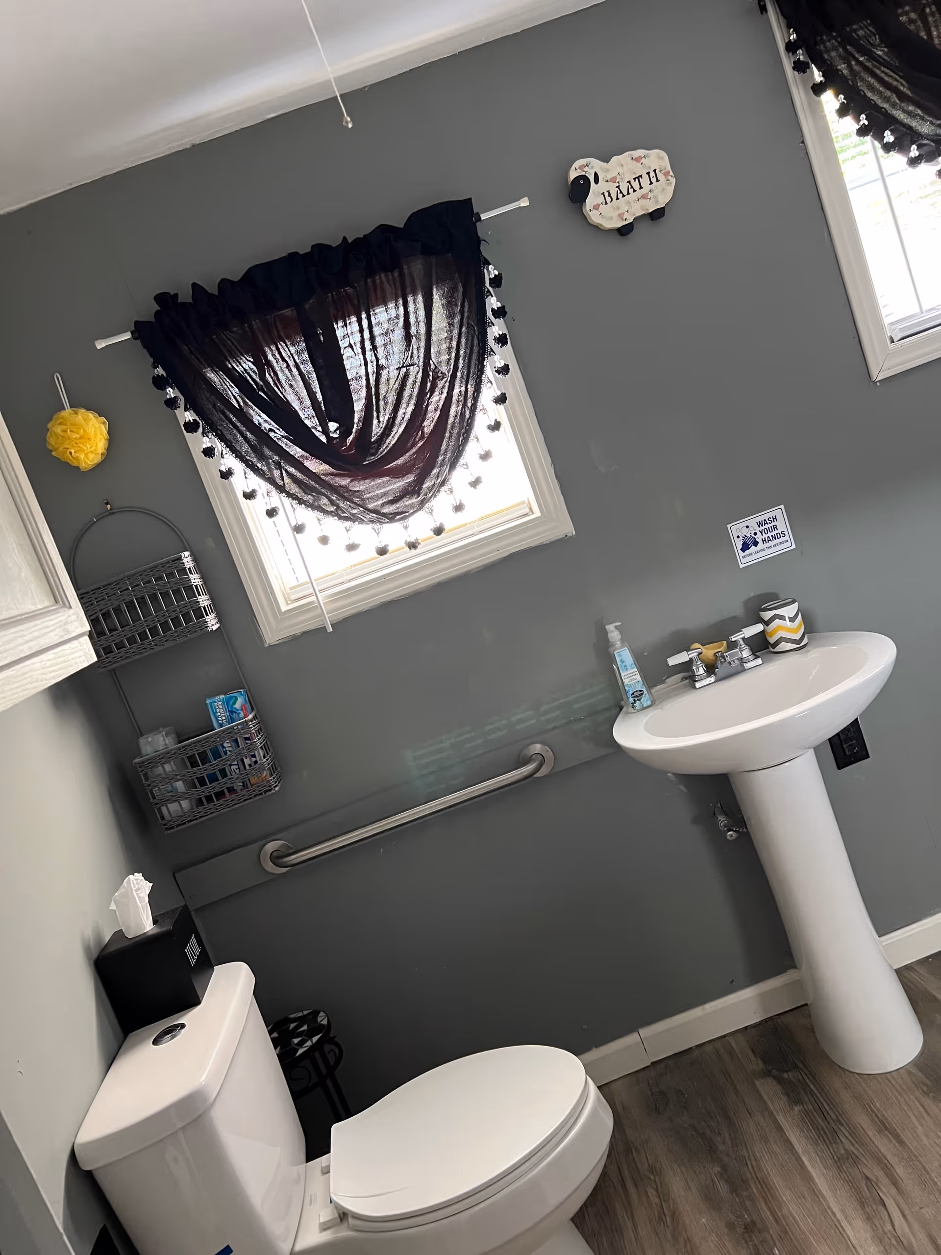 Small assisted living bathroom with pedestal sink, toilet, grab bar, and black sheer-curtained window.