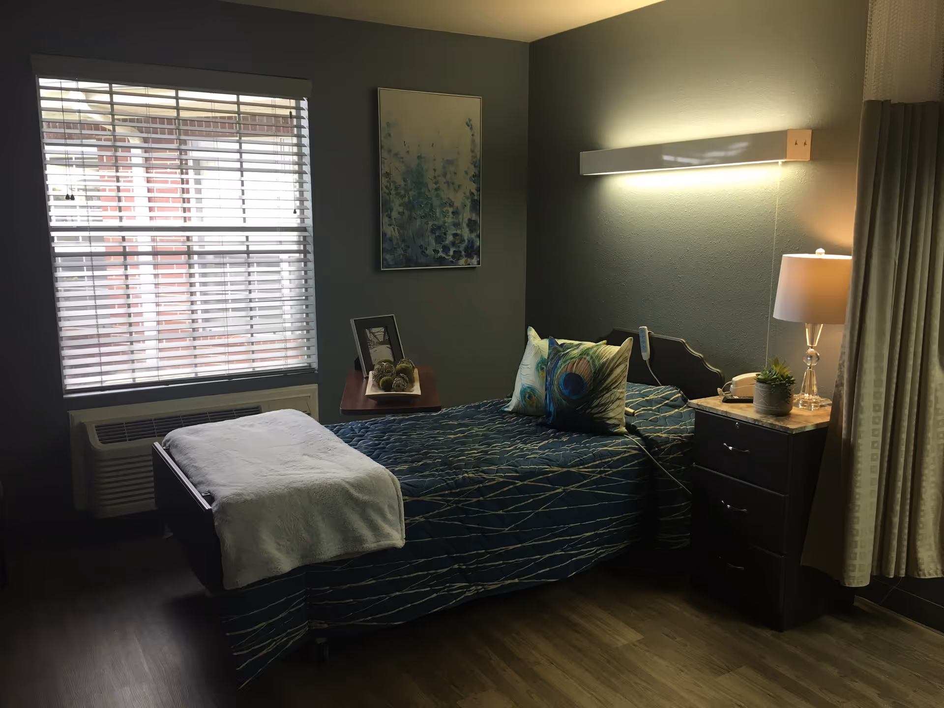 Bedroom with a single bed, blue quilt, pillows, nightstand, lamp, and window blinds.