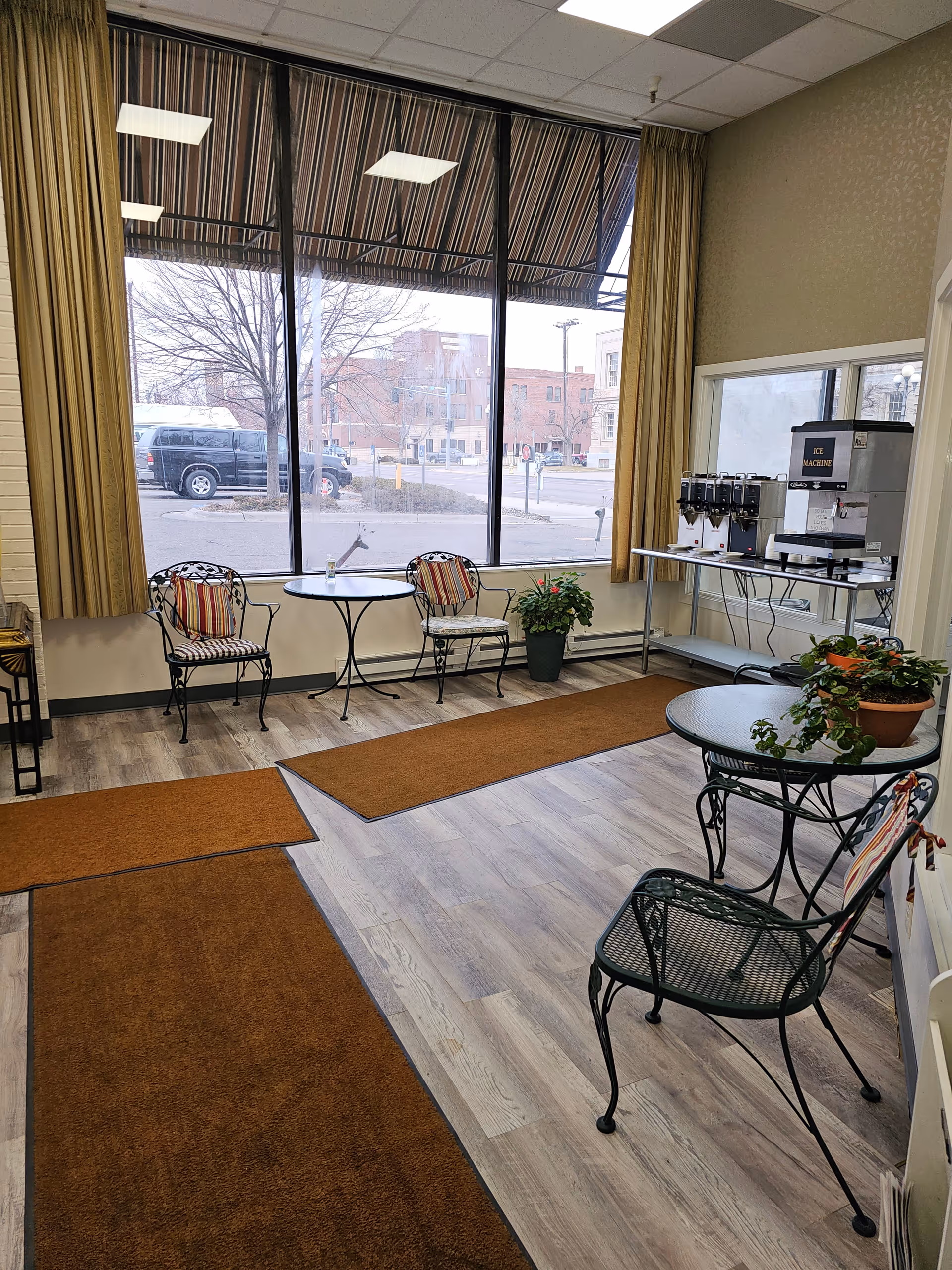 A bright interior common area with large windows showing an outside street view. The room has light wood flooring with brown rugs, metal chairs with cushions, and round tables. There is a beverage station with an ice machine and drink dispensers on a metal table against the wall. Potted plants are placed on the tables and floor.