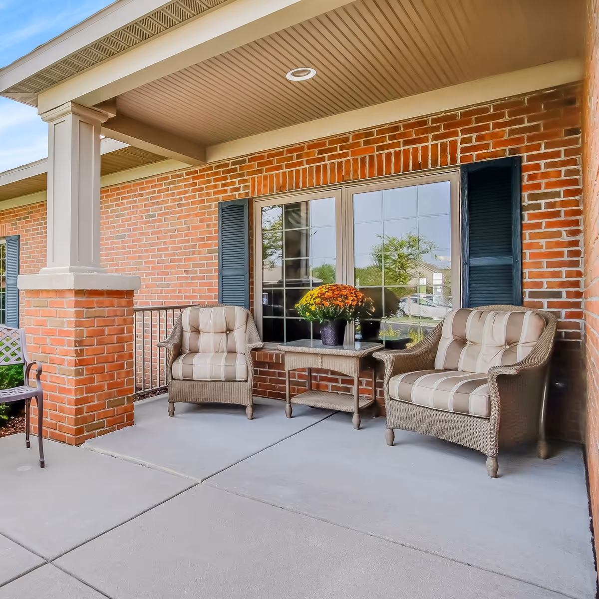 Covered brick front porch with two cushioned wicker chairs, a small table holding a pot of flowers, and windows with shutters.