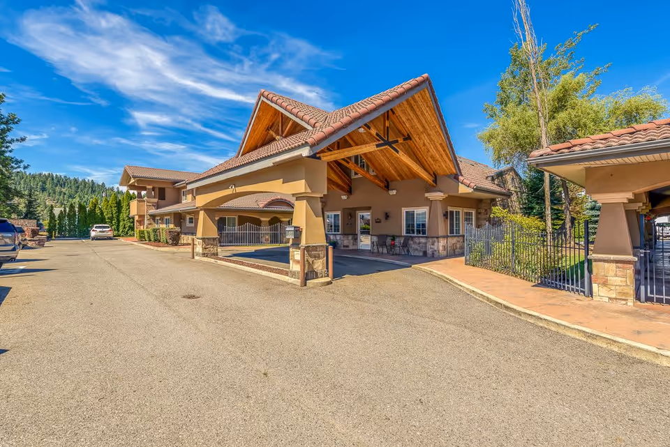 Exterior view of Pinehurst Senior Community building with a large covered entrance, stone and stucco facade, surrounded by trees and a clear blue sky.