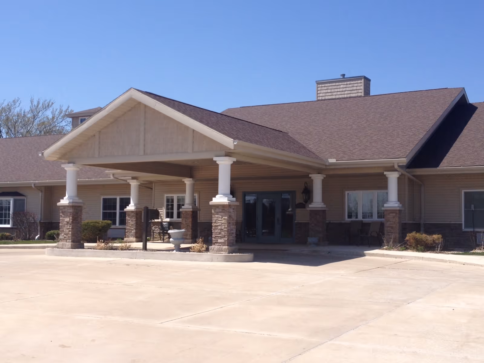 Exterior front view of a single-story building with a large covered entrance supported by white columns with stone bases, beige siding, and a brown shingled roof under a clear blue sky.