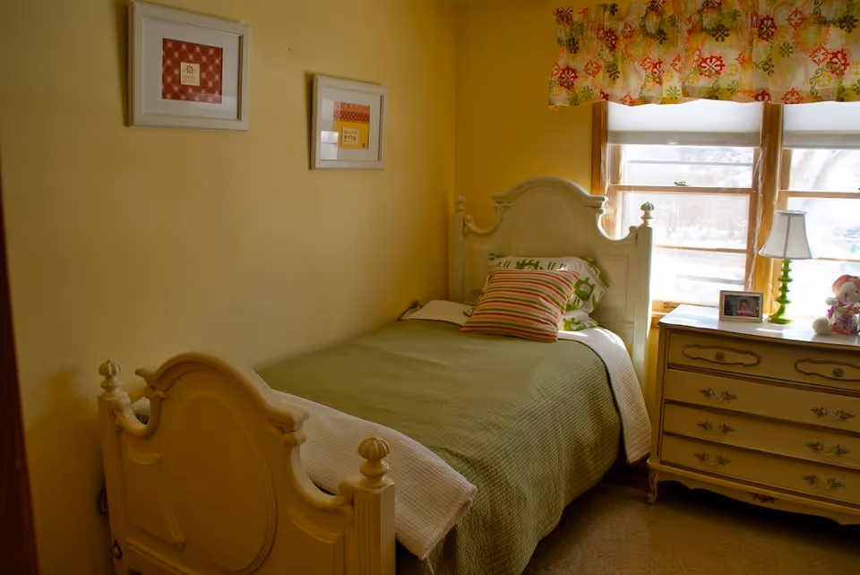 A cozy bedroom with a single white ornate bed featuring a green bedspread and striped pillow. Next to the bed is a vintage-style dresser with a green lamp, a framed photo, and a teddy bear. Two windows with floral valances let in natural light, and two framed pictures hang on the yellow wall.
