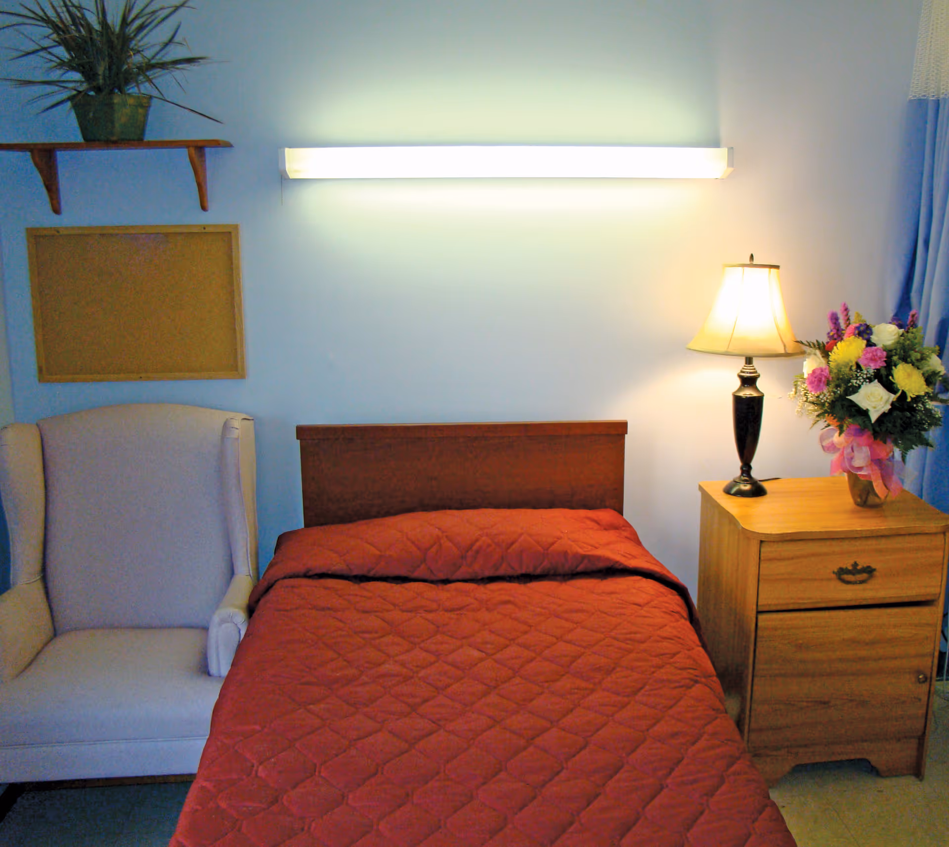 A small bedroom with a single bed covered in a red quilt. To the right of the bed is a wooden nightstand with a lamp and a vase of colorful flowers. To the left of the bed is a light-colored armchair. Above the bed is a long wall-mounted light fixture, and on the wall to the left is a small shelf with a potted plant and an empty corkboard.