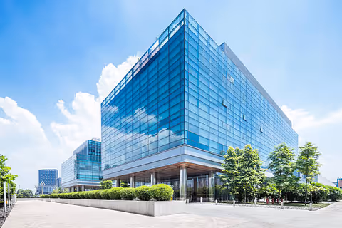 A large glass-fronted facility with blue reflections, surrounding trees, and a clear bright sky.