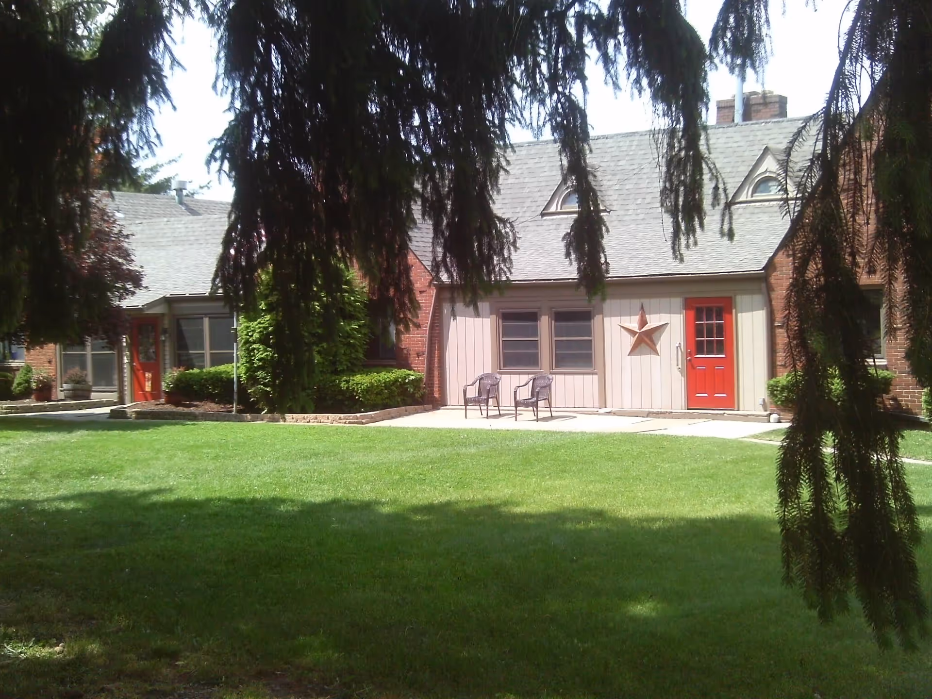 View of a senior living facility building with a well-maintained green lawn in front. The building has a gray roof, beige siding with a large star decoration, two red doors, and two chairs placed on the concrete patio. Large tree branches hang down in the foreground, partially shading the scene.