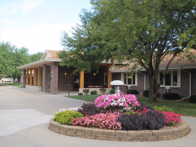 Front entrance of a brick senior living building with a circular flower bed of pink flowers and trees.