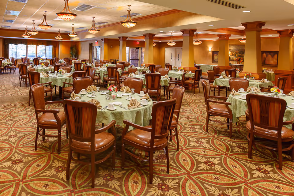 Spacious dining room with round tables set for meals, wooden chairs, chandeliers, and patterned carpet.
