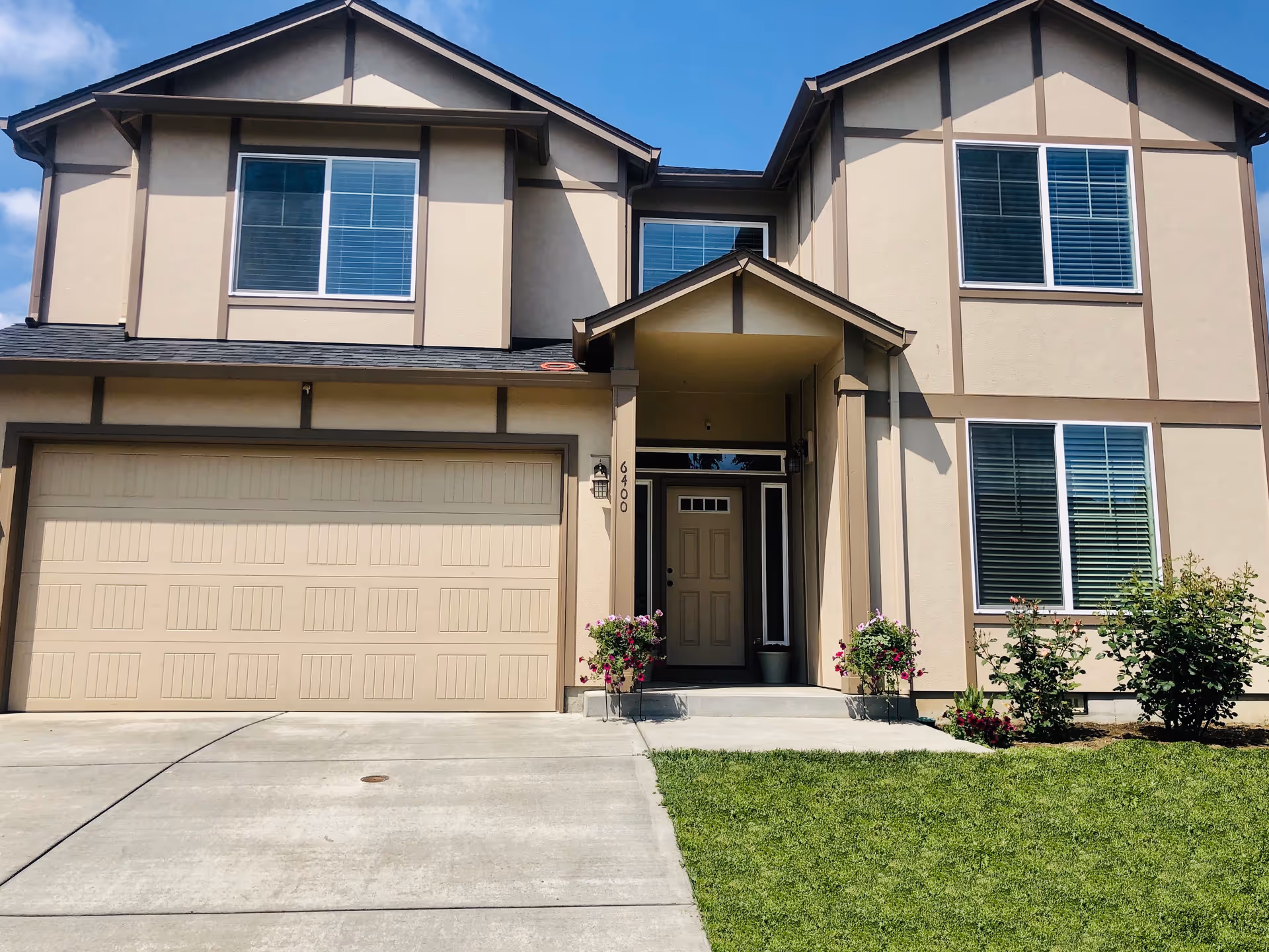 Front exterior of a two-story beige house with a double garage, central entrance, windows with blinds, and a small lawn with potted flowers.