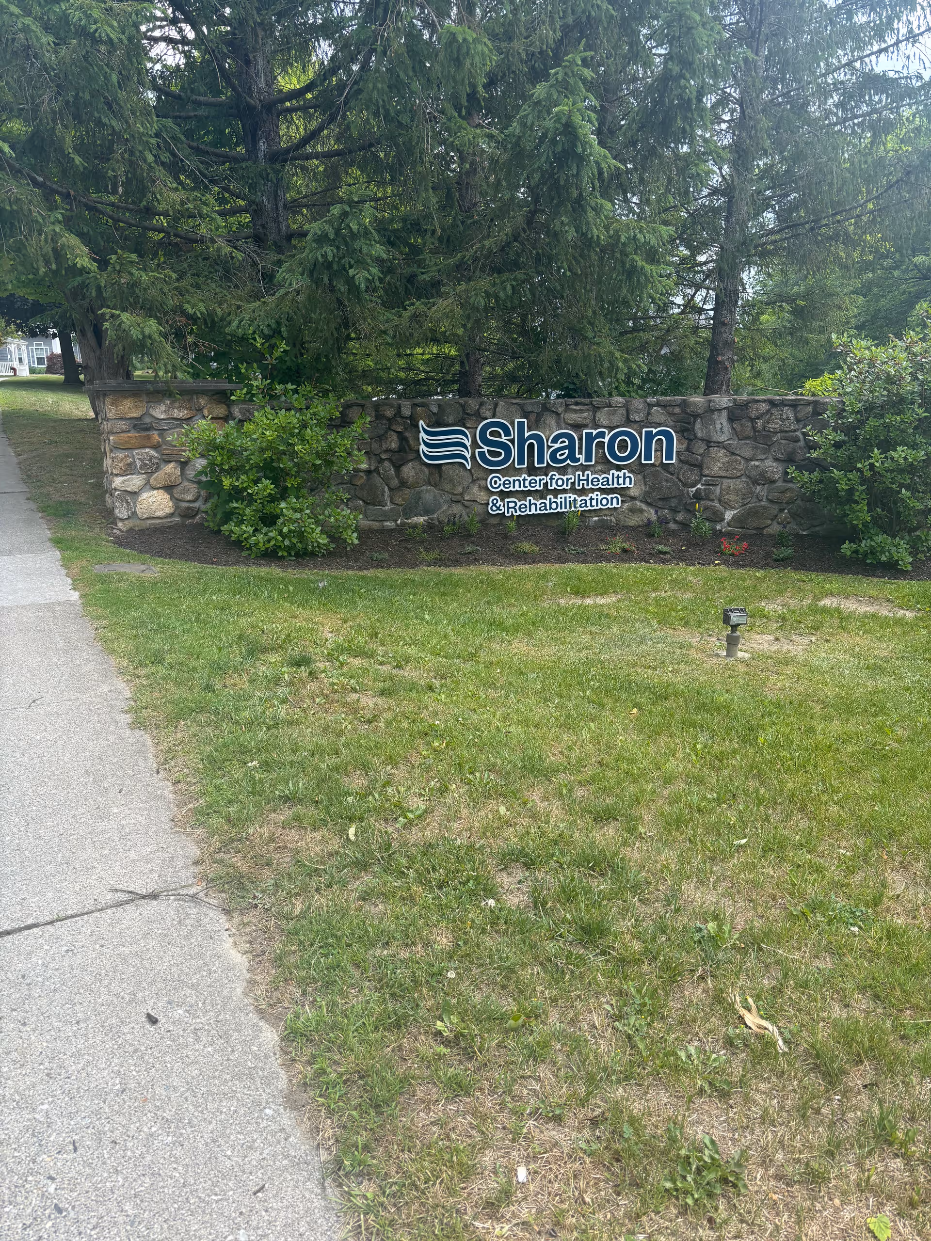 Stone wall sign reading Sharon Center for Health & Rehabilitation, surrounded by shrubs and trees.