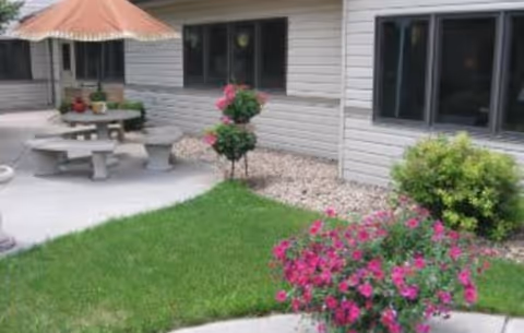 Outdoor patio beside a building with a round concrete picnic table, umbrella, and flowering plants.