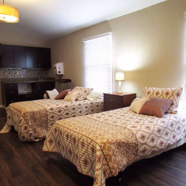 Two single beds with patterned bedspreads, a nightstand and lamp, and a small sink/cabinet area in a neutral-toned bedroom.