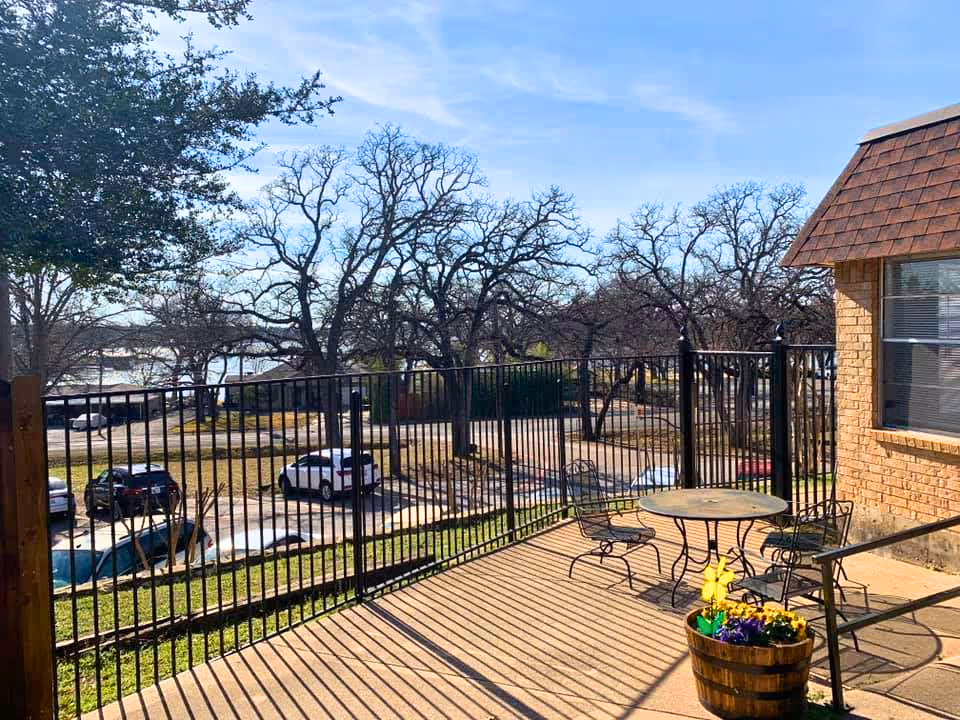 Sunlit outdoor patio with a metal table and chairs and a flower planter overlooking a fenced parking lot, trees, and a lake.
