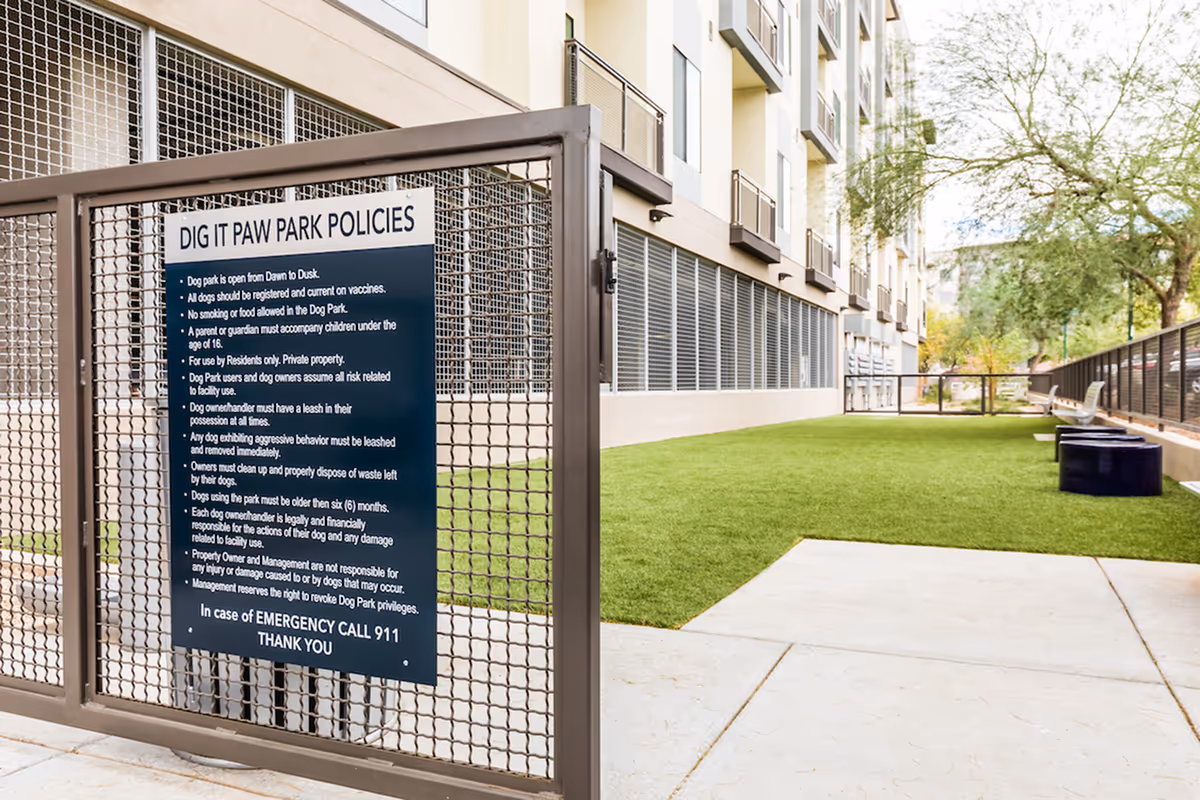 A fenced dog park area with a sign displaying Dig It Paw Park policies. The park has artificial grass, benches, and is adjacent to a multi-story residential building with balconies. Trees and a clear sky are visible in the background.