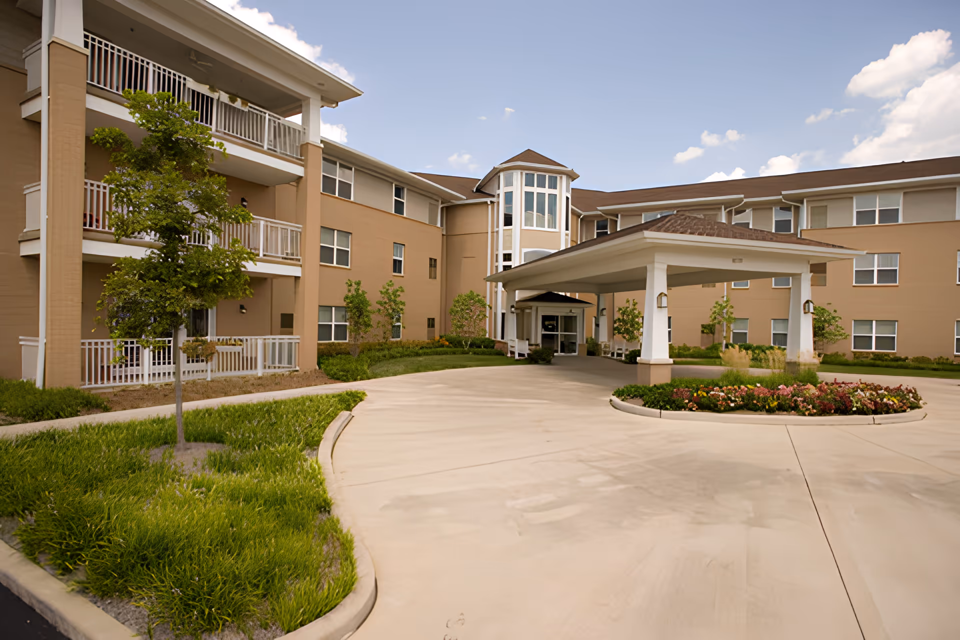 Front exterior of a multi-story assisted living building with balconies, a covered entrance/porte-cochere, and landscaped driveway.