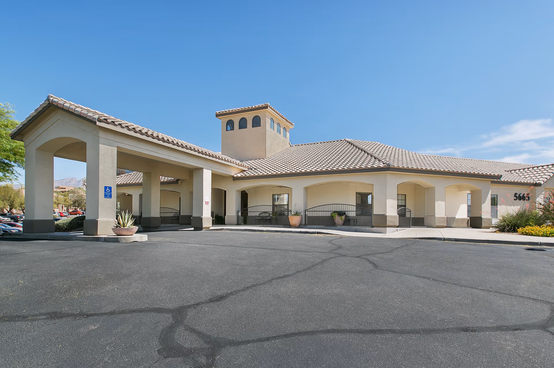Exterior view of a single-story building with a tiled roof and arched entryways under a clear blue sky. The building has a covered entrance with columns and a wheelchair accessible sign. There are potted plants near the entrance and a parking lot in front.