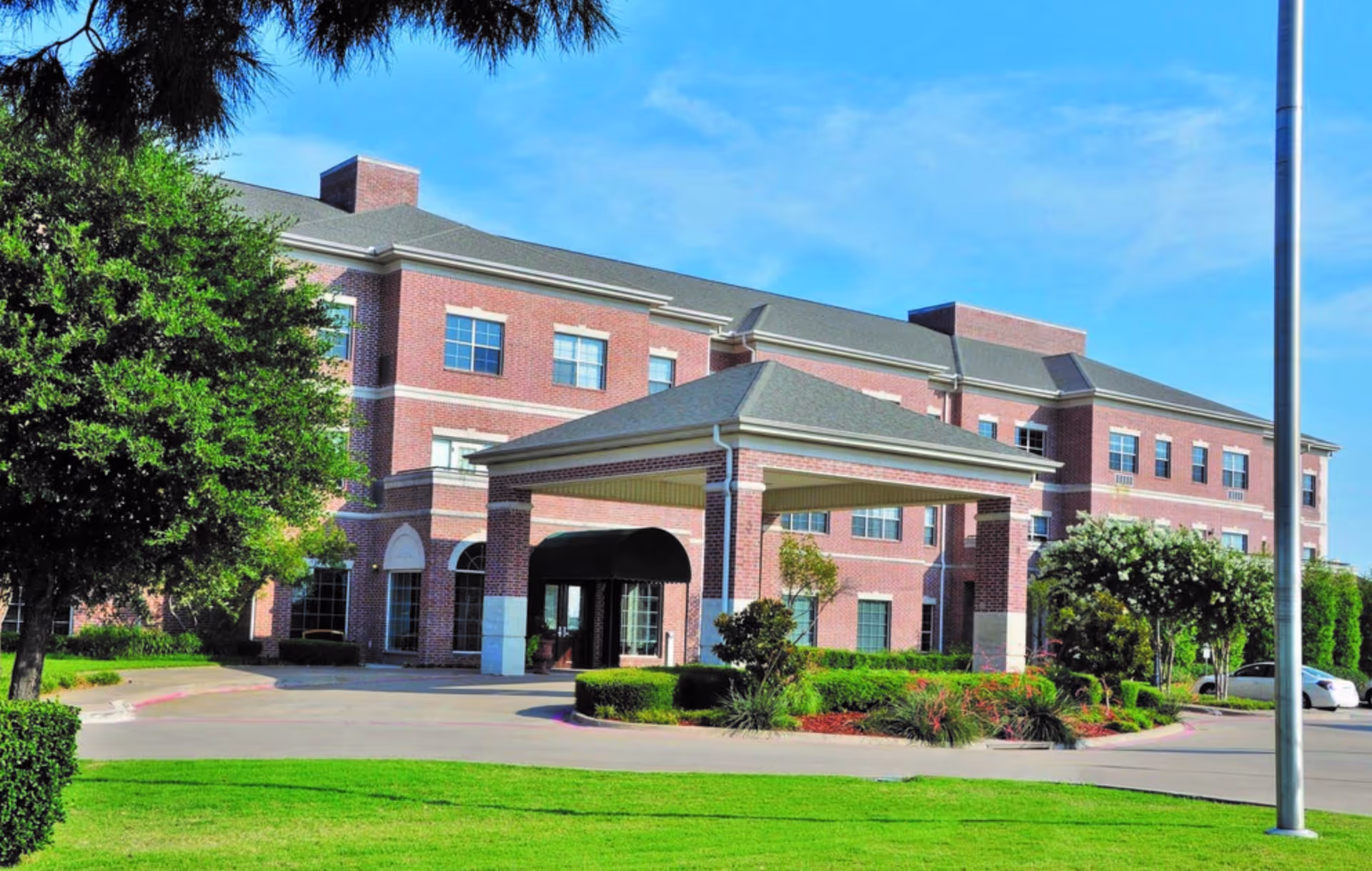 Front entrance of a three-story red brick senior living facility with a covered drop-off portico and landscaped grounds.