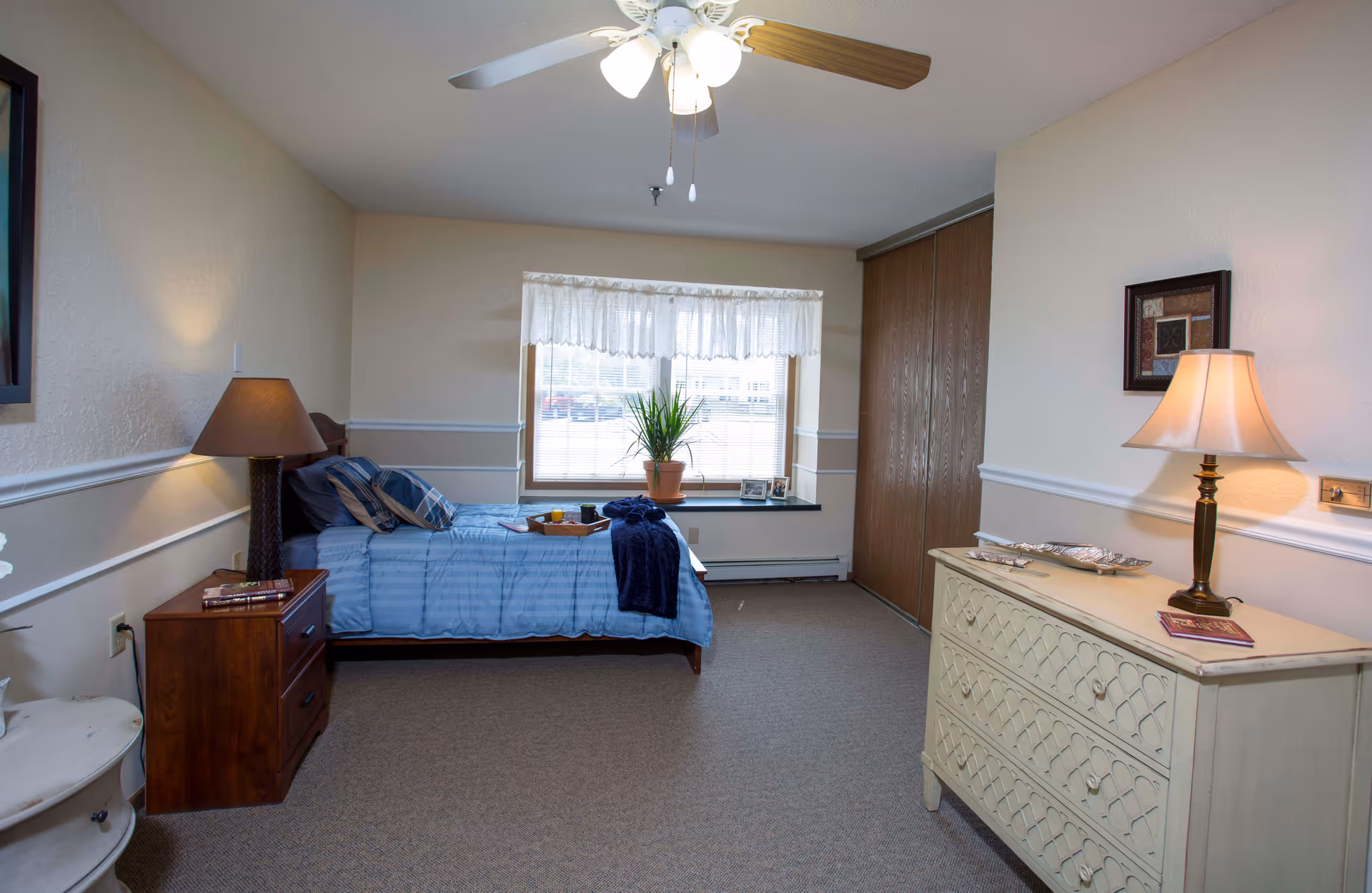 Senior living bedroom with blue bed, dresser, nightstand, window, and ceiling fan.