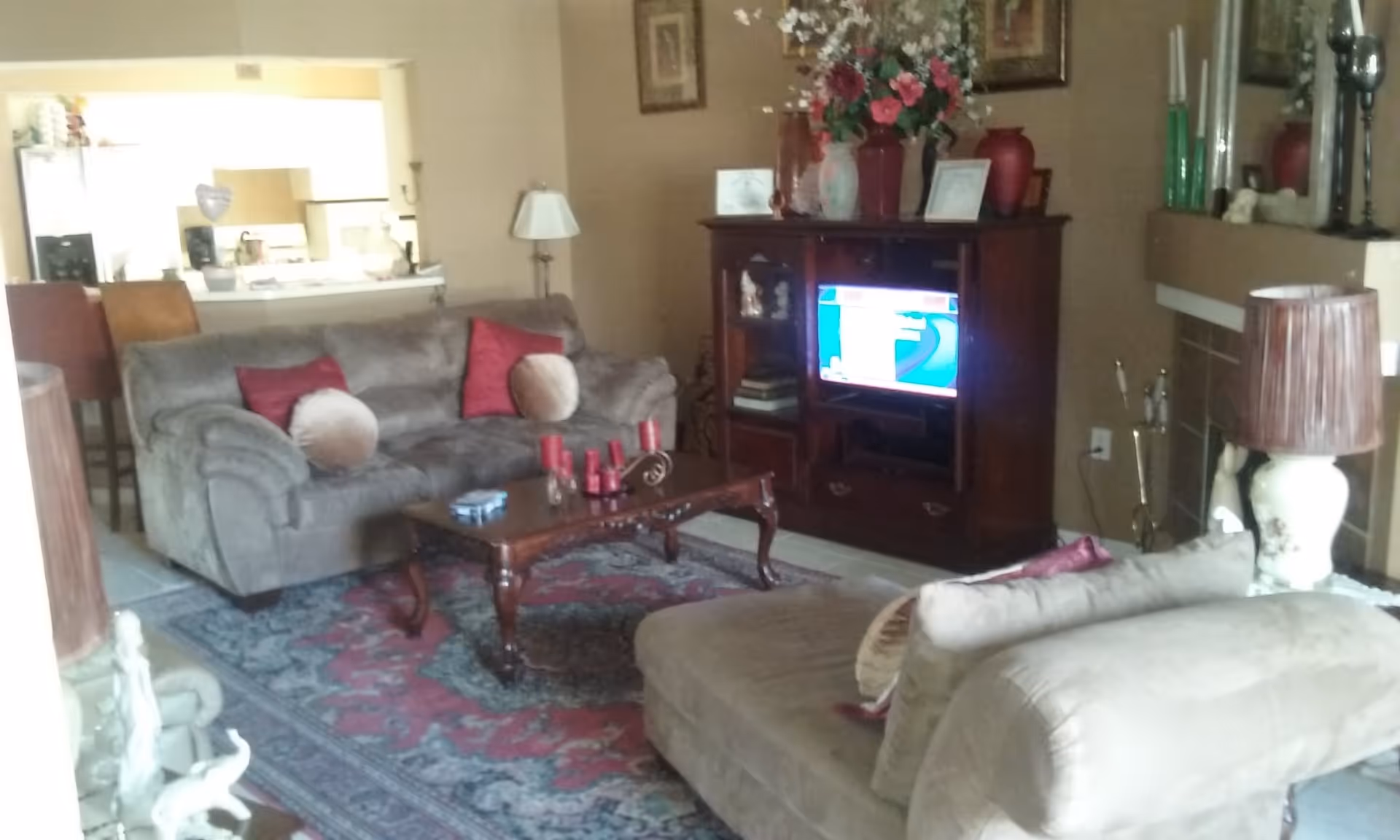 A cozy living room with two beige sofas adorned with round and square cushions, a wooden coffee table with red candles, a patterned area rug, a wooden TV cabinet with a television on, and various decorative items including vases and framed pictures. The room has a warm color scheme with beige walls and a fireplace on the right side.