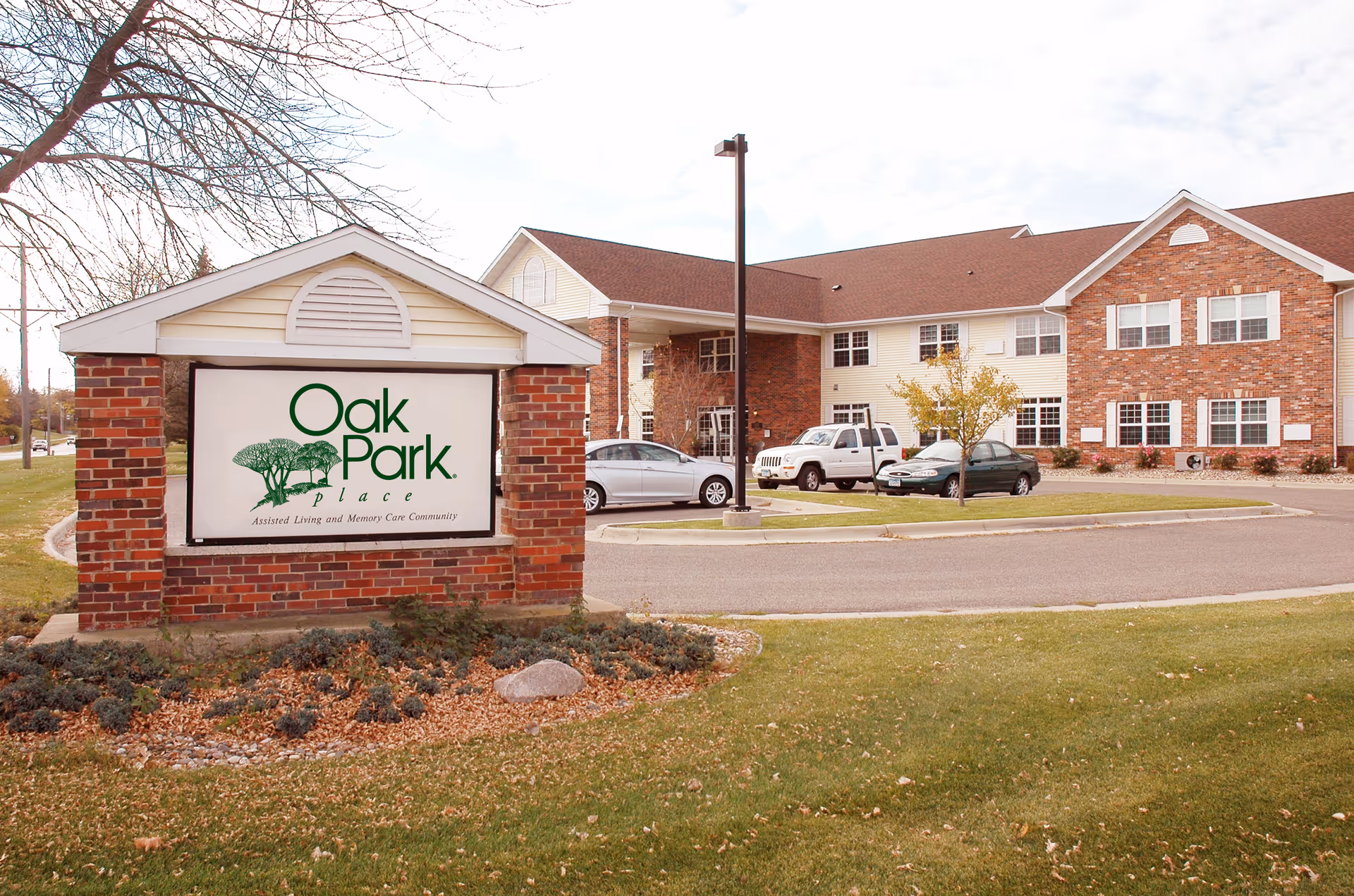 Exterior view of Oak Park Place Albert Lea assisted living and memory care community building with a brick and beige siding facade, parked cars, and a large sign in front displaying the facility name and logo.