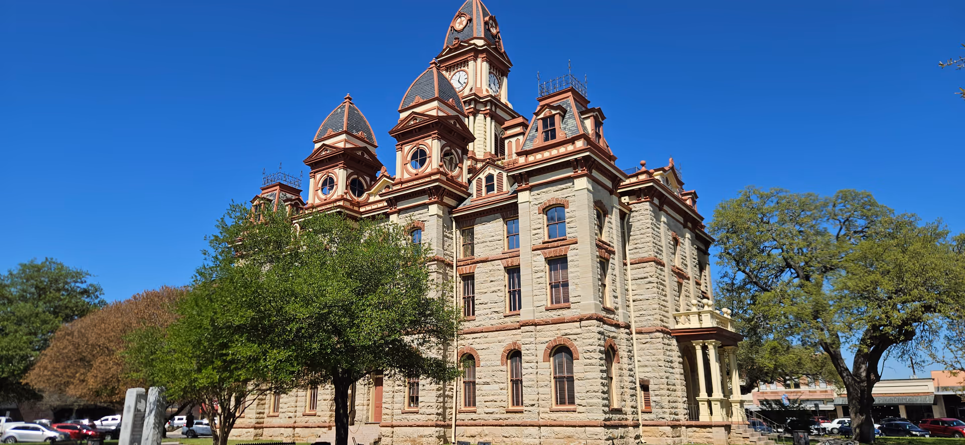 Historic stone building with towers, trees, and parked cars under a bright blue sky.