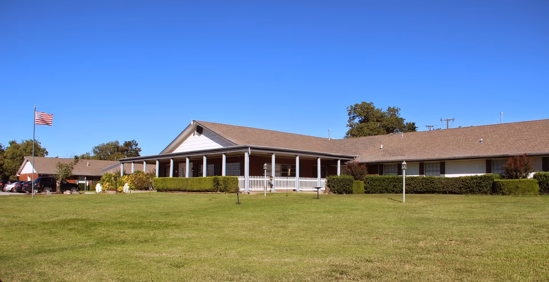 Single-story nursing home building with a large front porch, white picket fence, and well-maintained green lawn under a clear blue sky. An American flag is flying on a flagpole to the left, and several cars are parked near the building.