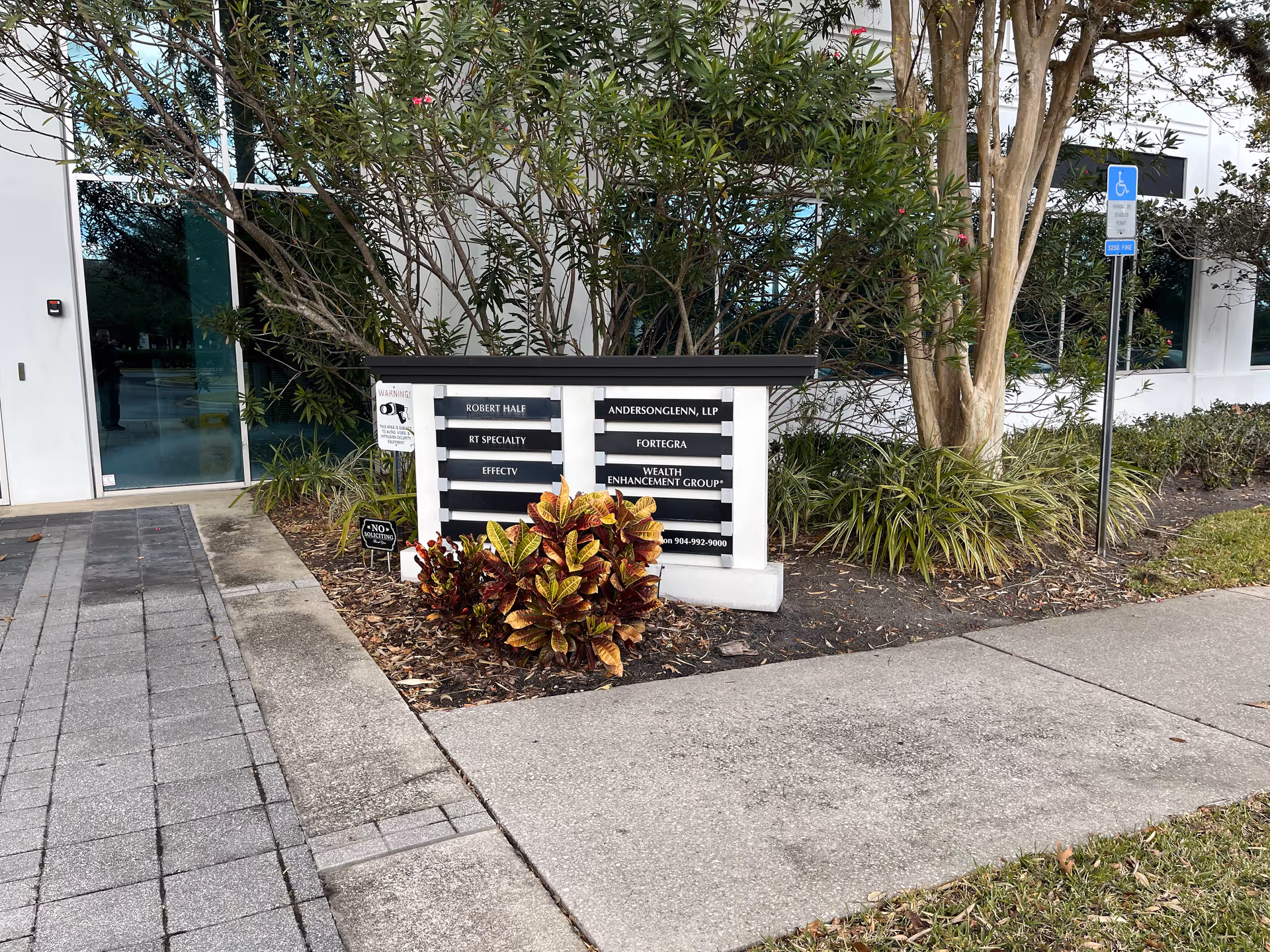 Exterior facility entrance with signboard, landscaping, and a wheelchair parking sign.