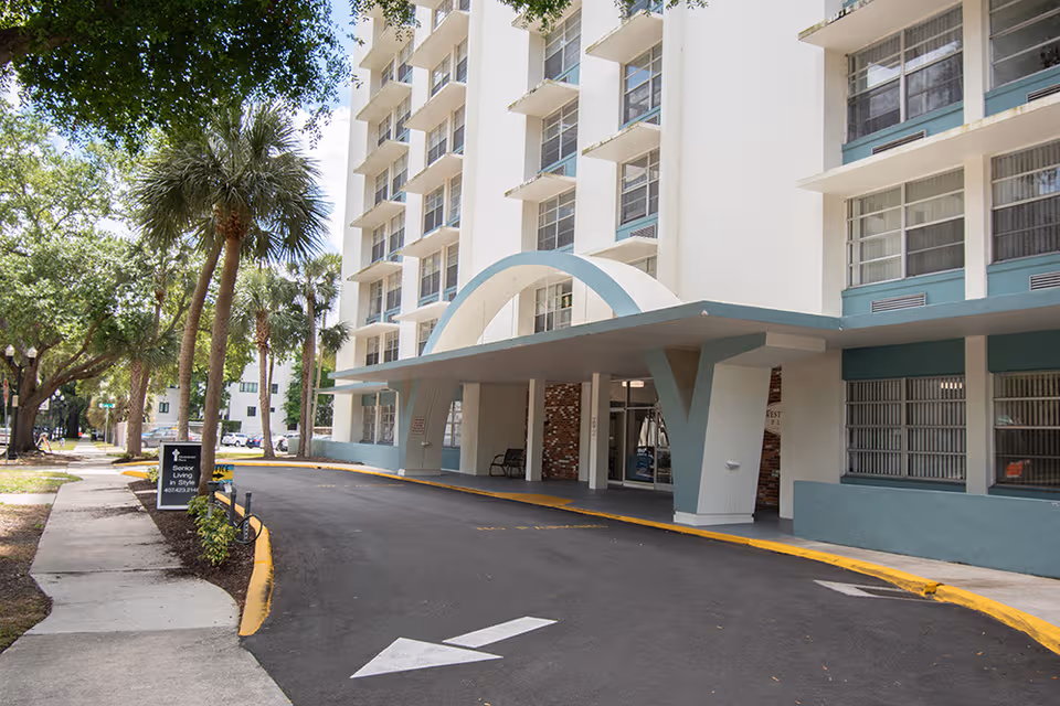White multi-story senior living building with curved entrance canopy and palm trees.