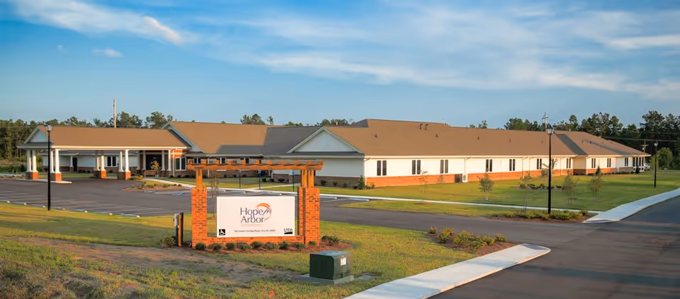 Exterior view of Hope Arbor Of Loris, a single-story senior living facility with a large parking area, green lawn, and a sign displaying the facility's name in front.