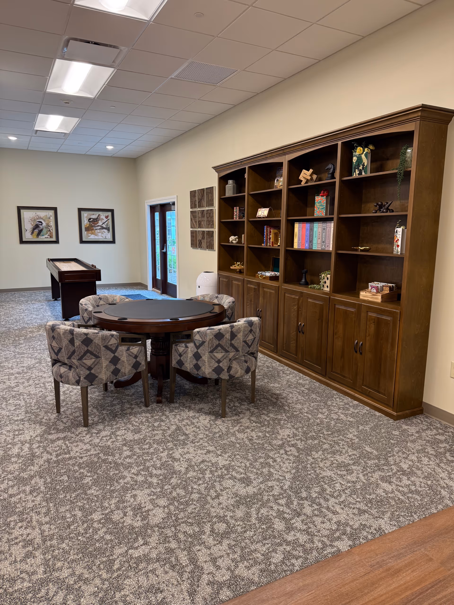 A spacious senior living lounge with a round table, patterned chairs, and a tall wooden bookshelf.
