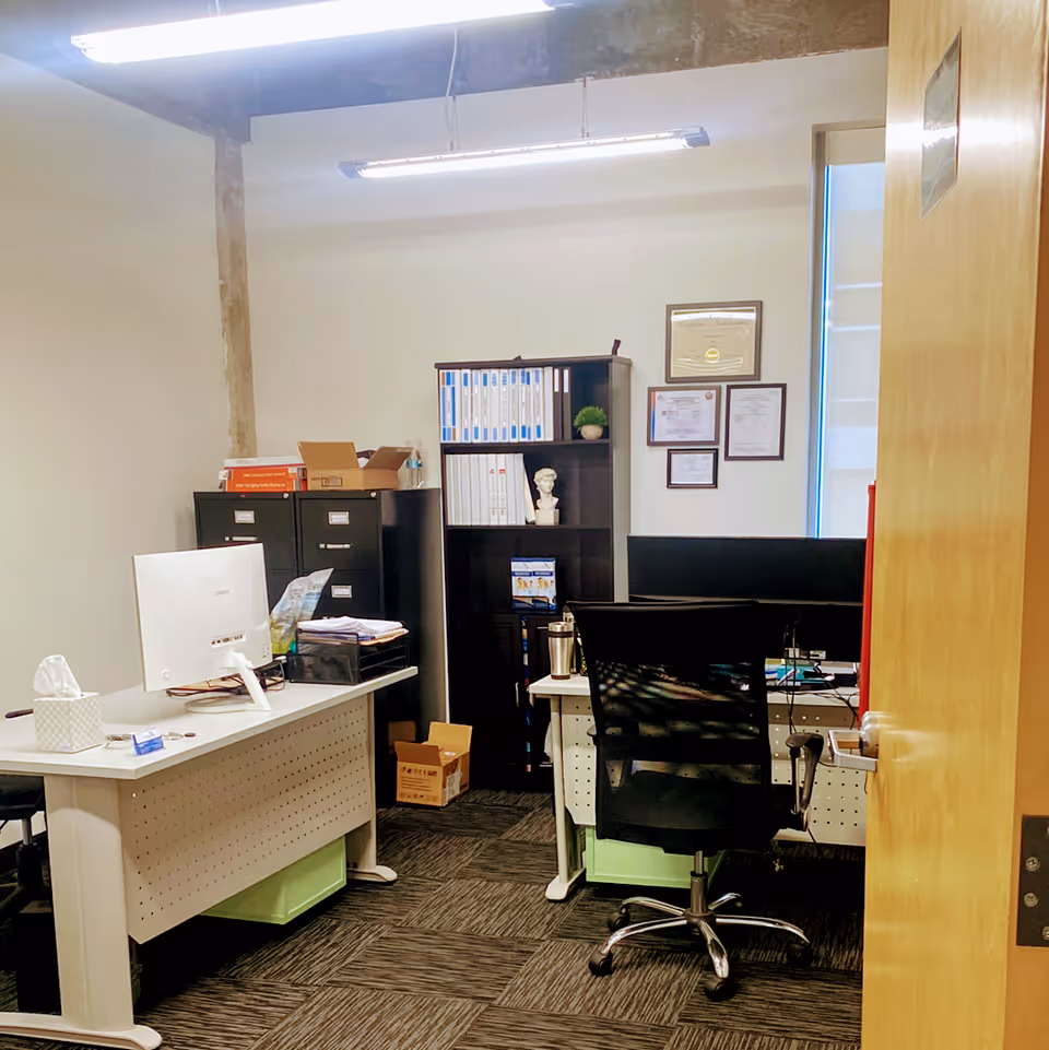Office workspace with desks, computers, filing cabinets, and certificates on the wall.