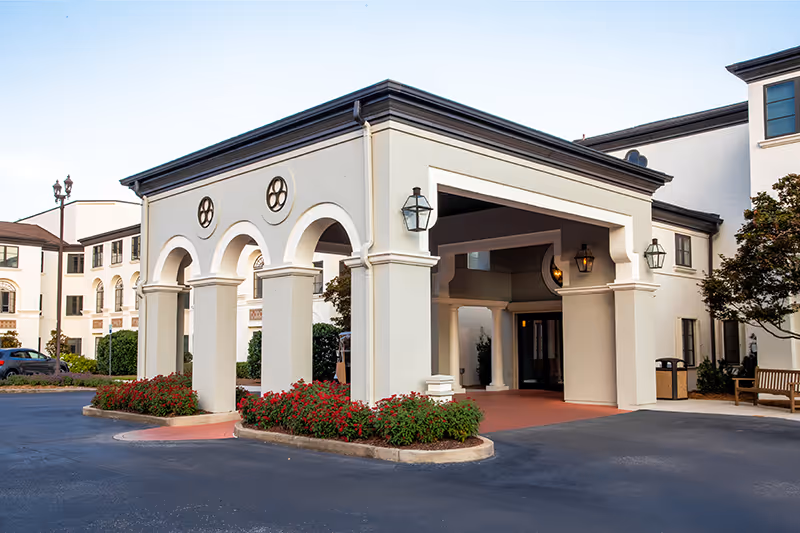 Entrance of a senior living facility with a covered drop-off area supported by columns and arches, surrounded by flower beds and a paved driveway. The building is white with black trim and has outdoor lantern-style lights mounted on the walls.