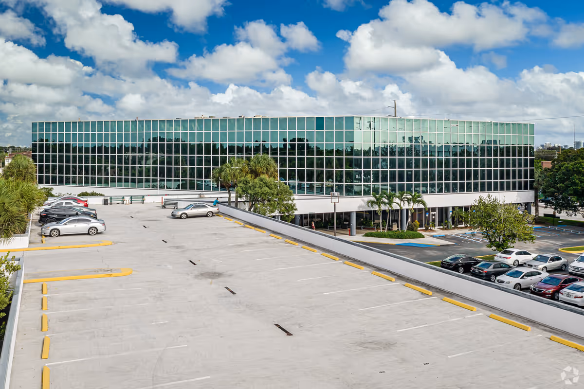 View of a modern glass-front building with a large parking lot in the foreground.
