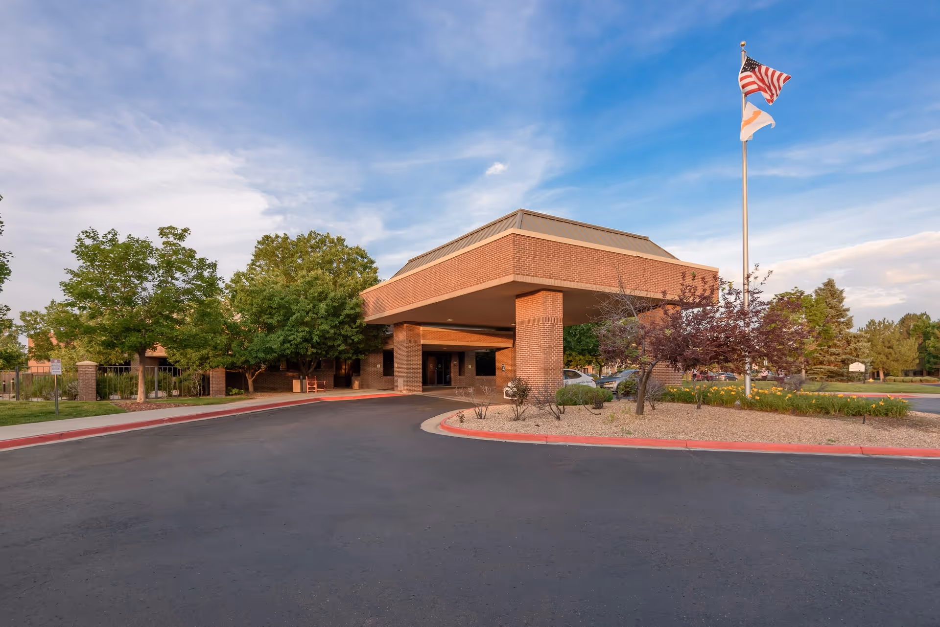 Exterior view of a senior living facility entrance with a covered drop-off area, surrounded by trees and landscaping. An American flag and another flag are flying on a flagpole near the entrance under a partly cloudy sky.