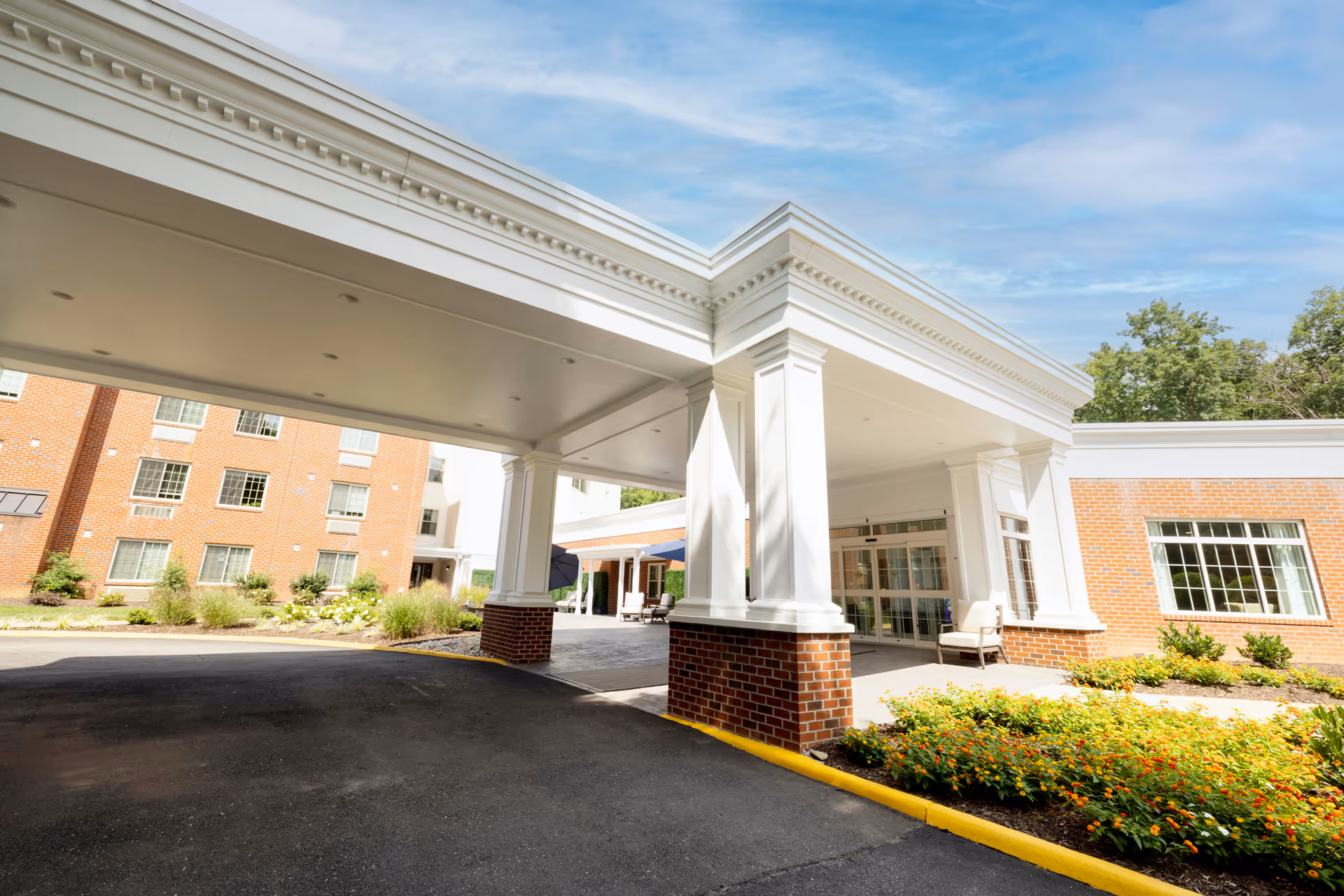 Entrance area of a senior living facility with a covered drop-off zone supported by white columns with brick bases. The building has large windows and is surrounded by landscaped flower beds and greenery under a partly cloudy sky.
