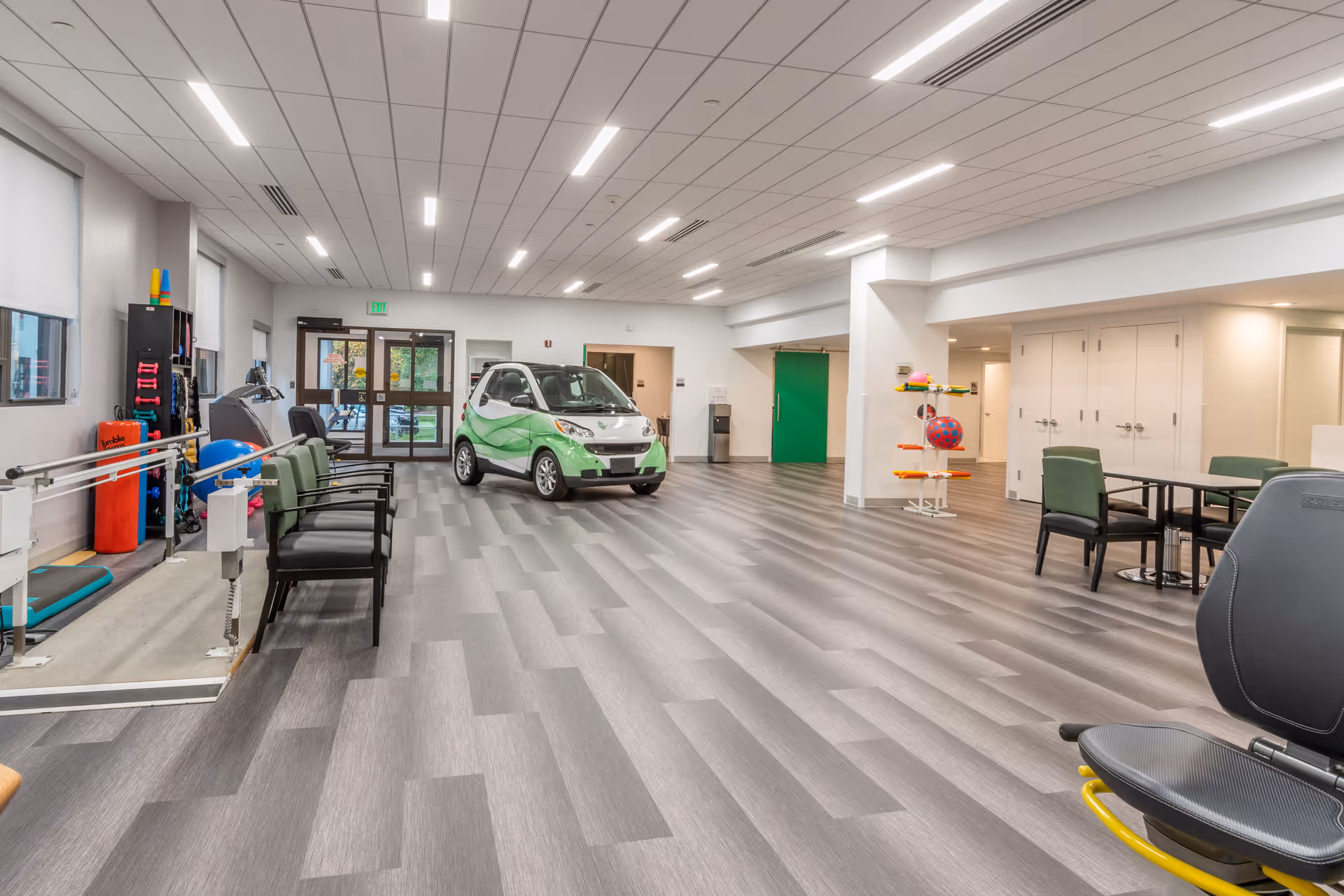 A bright, spacious indoor rehabilitation room with exercise equipment, seating, and a small green-and-white therapy car.