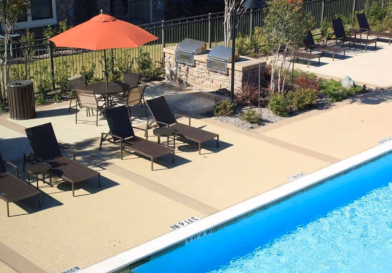 Poolside patio area with lounge chairs, a table under an orange umbrella, built-in grills, and a swimming pool.