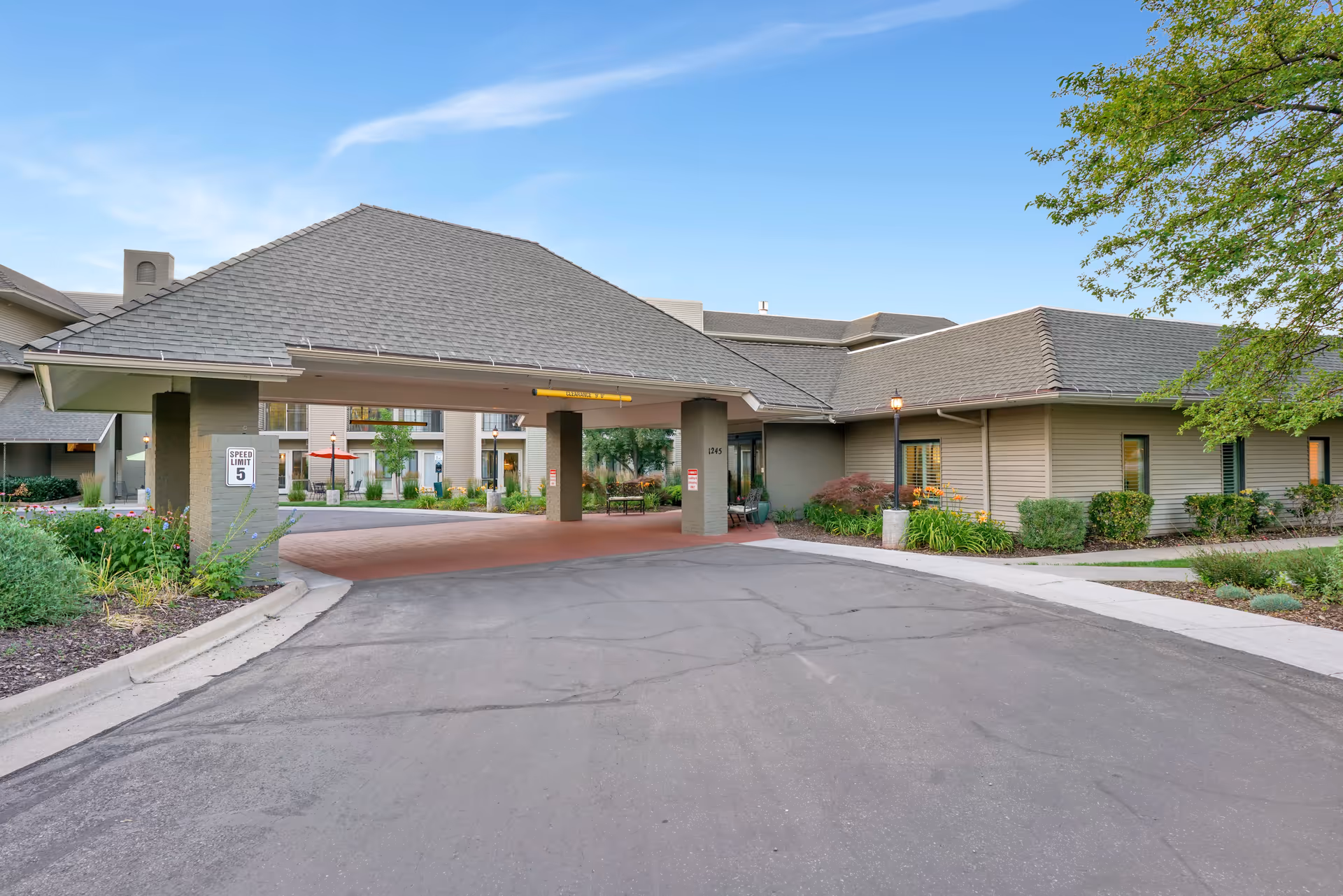 Entrance to a senior living facility with a covered drop-off area, surrounded by landscaped greenery and a clear blue sky.