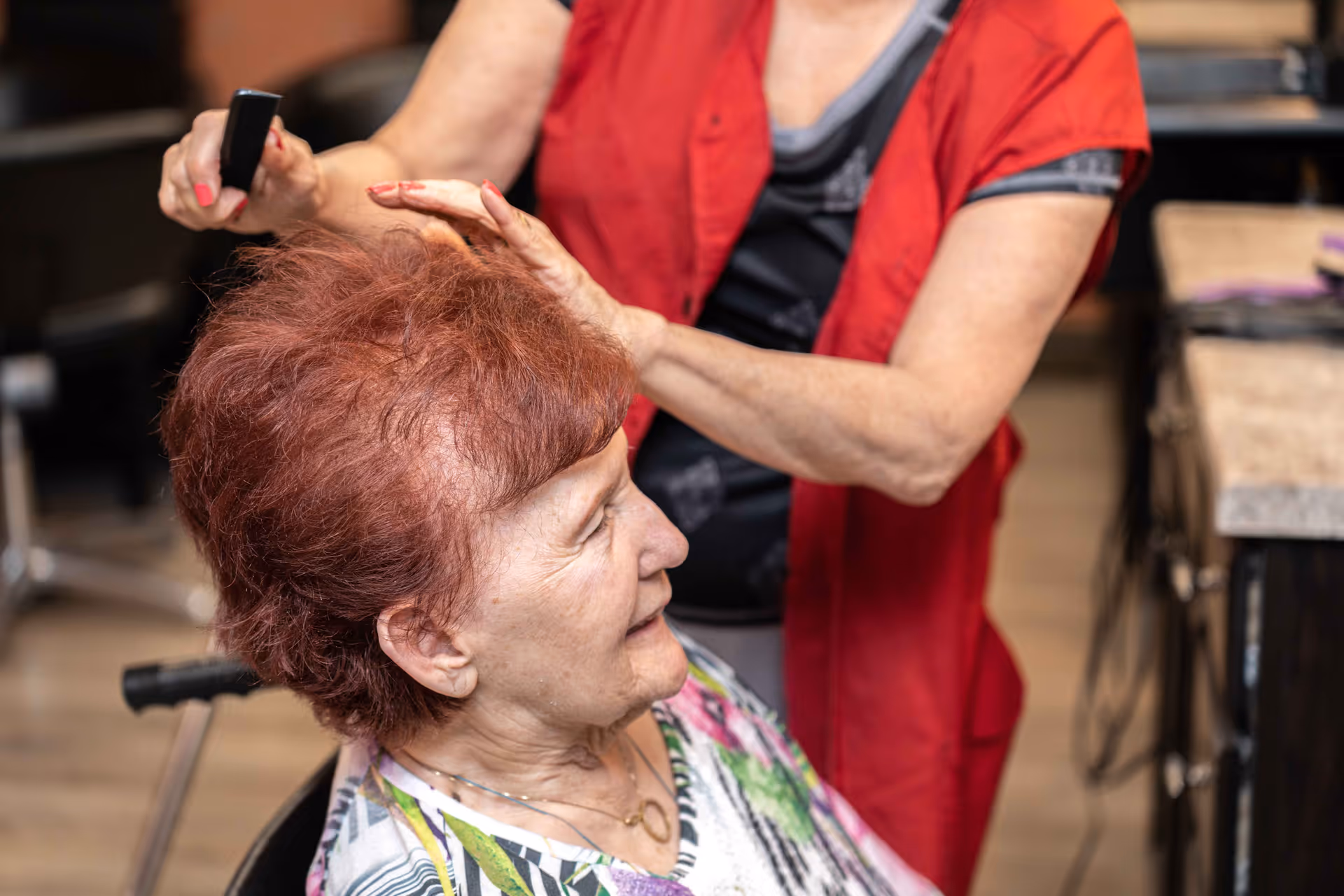 An elderly woman with short red hair is sitting in a chair while another person, wearing a red vest, styles her hair with a comb in a salon-like setting.